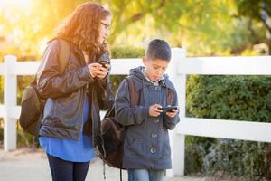hermano y hermana hispanos con mochilas caminando enviando mensajes de texto en teléfonos celulares foto