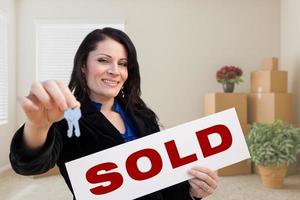 Hispanic Female Real Estate Agent with Sold Sign and Keys in Room with Moving Boxes. photo