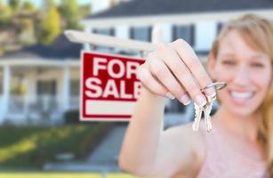 Excited Woman Holding House Keys and For Sale Real Estate Sign in Front of Nice New Home. photo