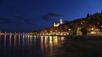 menton förbi natt från de strand. cote d'azur i provence, Frankrike. medelhavs hav. helgon michel basilika kyrka på kväll Timelapse video