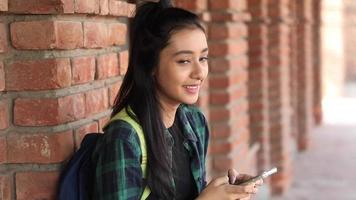 Video clip of a smiling Asian female college girl using her mobile phone near the college campus wall.
