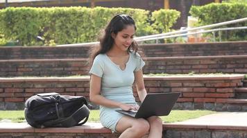 Stock video of an Indian teen girl sitting on a college campus and typing on a laptop.