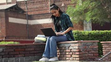 video de una universitaria india escribiendo en una computadora portátil mientras está sentada afuera en el área del campus universitario.