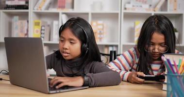 retrato de dos estudiantes asiáticas sentadas en el escritorio en casa. niña de pelo corto aprendiendo en línea a través de una computadora portátil y gafas de niña jugando en un teléfono inteligente. concepto de educación