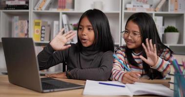 portret van twee Aziatisch leerling meisjes zittend Bij bureau en golvend hand- via laptop Bij huis. meisje kort haar- en meisje bril aan het leren online via computer. jong vrouw schrijven een boek. onderwijs concept. video