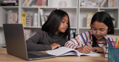 retrato de dos estudiantes asiáticas sentadas en el escritorio en casa. niña de pelo corto y gafas de niña aprendiendo en línea a través de una computadora portátil. mujer joven escribiendo un libro y escribiendo una computadora. concepto de educación