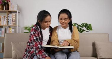 gelukkig Aziatisch tweeling meisjes lezing een boek en zittend Aan bankstel in leven kamer. werkzaamheid binnen- voor tiener in vakantie. opleiding, levensstijl en hobby concept.