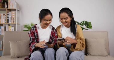 meninas gêmeas asiáticas felizes assistindo smartphone enquanto está sentado no sofá na sala de estar. mulheres engraçadas jogando jogo no celular. conceito de hobby, tecnologia e estilo de vida. video