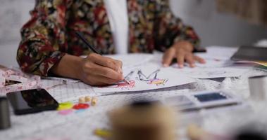 la mano ravvicinata della stilista donna asiatica disegna uno schizzo di abbigliamento femminile mentre è seduto in studio. startup piccola imprenditrice è in procinto di creare una nuova collezione di vestiti. video