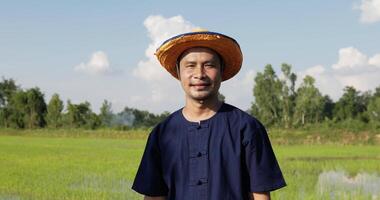 cámara lenta, retrato en la cabeza de un granjero, retrato de un adulto de mediana edad con camisa azul y sombrero de paja parado y mirando el campo de arroz de la cámara en el fondo y soleado video