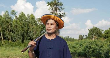 cámara lenta, retrata al agricultor de mediana edad con camisa y sombrero de paja llevando equipo en el hombro y caminando en los campos de arroz video