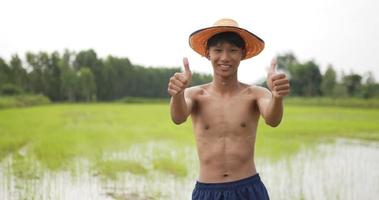 Front view, close up slow motion shot of male farmer, Portrait Young adult topless and wearing hat standing in rice field with his arms folded, smiling and looking at the camera and thump up video