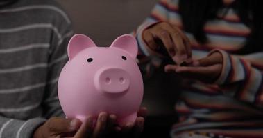 Closeup of Two hand girls putting coins into piggy bank while sitting on floor in living room. Young female holding piggy bank. Financial, saving money and investment concept.