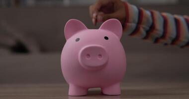 Closeup of Two hand girls putting coins into piggy bank while sitting on floor in living room. Front view piggy bank on table. Financial, saving money and investment concept. video