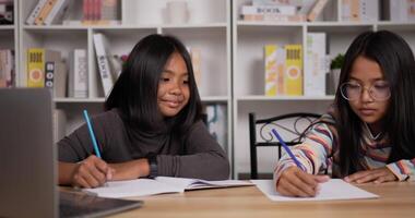 retrato de duas estudantes asiáticas sentadas na mesa em casa. menina de cabelo curto e óculos de menina aprendendo online via laptop. jovem fêmea escrevendo um livro. conceito de educação. video