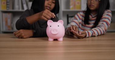 closeup de duas lindas garotas asiáticas economizando dinheiro e colocando moedas no cofrinho enquanto está sentado na mesa em casa. conceito financeiro e de investimento video