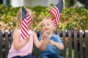 hermana joven y hermano ondeando banderas americanas en el banco en el parque foto