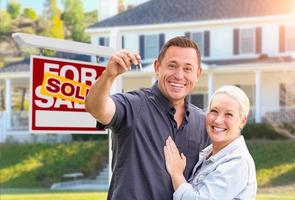 pareja feliz con nuevas llaves de casa frente al letrero de bienes raíces vendidos y hermosa casa foto