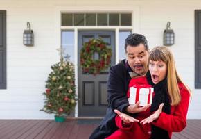 Young Mixed Race Couple Exchanging Gift On Front Porch of House with Christmas Decorations photo