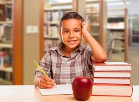 niño hispano con libros, manzana, lápiz y papel en la biblioteca foto