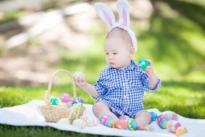 bebé de raza mixta chino y caucásico afuera usando orejas de conejo jugando con huevos de pascua foto