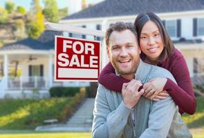 Mixed Race Caucasian and Chinese Couple In Front of For Sale Real Estate Sign and House. photo