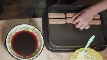 las manos de las mujeres hacen pastel de tiramisú. Remoje las galletas en el café y extiéndalas uniformemente. video