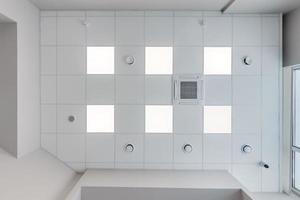 cassette stretched or suspended ceiling with square halogen spots lamps and drywall construction with fire alarm and ventilation in empty room in house or office. Looking up view photo