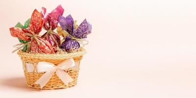 huevos de pascua envueltos en papel de color en una cesta decorada con lazo sobre fondo rosa pastel. bandera. foto