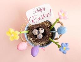Nest of quail eggs, decorative eggs, flowers, note Happy Easter on pastel pink. Top view. photo