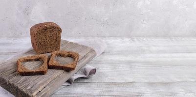 Loaf of rye bread and two slices with carved holes of heart shape in them on old wooden Board on gray background. photo