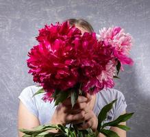 imagen festiva creativa con una joven sin rostro con una camiseta gris sobre gris sosteniendo peonías rosas en sus manos. estilo moderno sin rostro. imagen irreconocible. día de la madre o fiesta de la mujer. foto