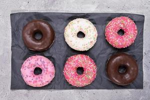 Six traditional doughnuts with multicolored glaze neatly laid out on black wrapping paper on grey concrete background. photo