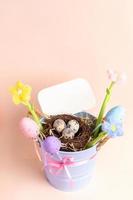 Blue bucket with nest of quail eggs, decorative eggs, flowers, empty note on pastel pink. Mock up. photo