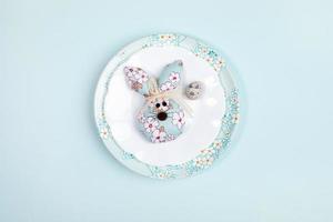 Easter festive minimal layout with one quail egg and one DIY textile Easter bunny on plates on light blue backdrop. photo