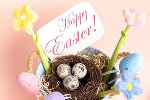 tarjeta de pascua con nido de huevos de codorniz, huevos decorativos, flores, nota feliz pascua de cerca en rosa. foto
