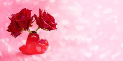Close-up three red roses in red vase shaped heart on pink bokeh background with hearts. Valentines day or mothers day festive banner. photo