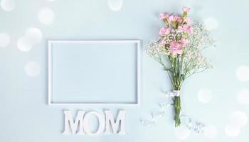 Empty frame, bouquet of carnation and gypsophila with ribbon, letters MOM on light blue. Top view. photo