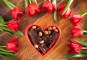 Red heart shaped bowl with chocolate and red tulips lying in around, on diagonal wooden background. photo