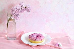 bodegón con ramita lila, donut en plato figurado y pequeños malvaviscos en forma de corazón sobre tela rosa sobre fondo rosa. foto