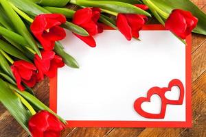 hoja blanca en blanco, marco rojo, tulipanes rojos y dos corazones rojos sobre fondo de madera diagonal. foto