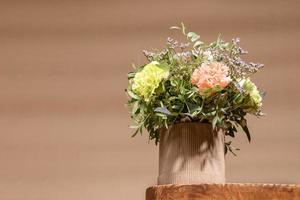 Ecological composition with flowers bouquet in DIY cardboard vase standing on old wooden table on beige with shadows. photo