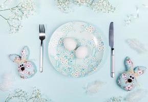 Easter table setting with white eggs on plate with floral decor, Easter bunnies and white flowers on pastel blue.