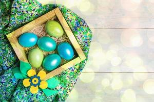 DIY Easter decor. Colored eggs in square cardboard box with flower on green floral fabric on wooden backdrop with bokeh. photo