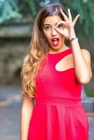 Young beautiful girl with red dress holds the circle with her fingers to the eye photo