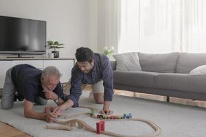 Happy caucasian old father and adult son family playing train toy together on holiday with feeling childhood nostalgic in cozy living room house background. Light effect. photo