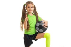 little girl in sports uniform with soccer ball in hands shows thumbs up photo