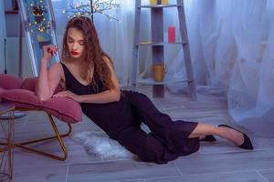 horizontal photo of Glamour woman with red lips in black overalls sits on the floor at home