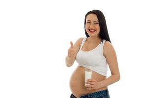 beautiful young pregnant brunette woman touching her belly and drinks milks showing thumbs up isolated on white background photo