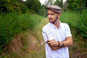 Elegant man in hat holds sunglasses and looking away at the green garden photo